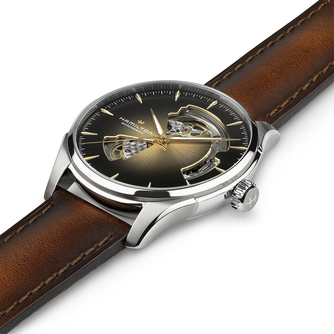 The Hamilton Jazzmaster Open Heart Auto 40mm Watch features a brown leather strap, silver case, and dark gradient cutout dial revealing its inner mechanism. “Hamilton” is displayed on the dial.