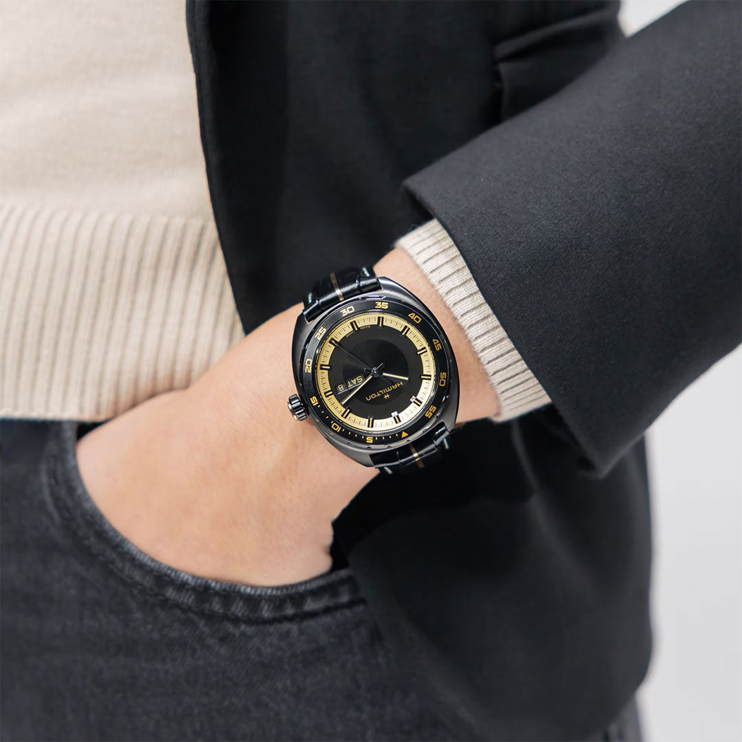 Wearing a Hamilton American Classic Pan Europ Resident Evil Requiem Limited Edition Auto 42mm watch with a black and gold case and crocodile-pattern leather strap, this person pairs it with dark jeans, a black blazer, and a light beige sweater.