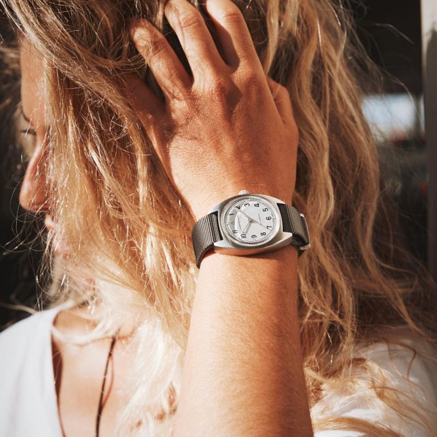 A person with blonde, wavy hair wearing a white shirt is holding their hair back, showcasing a Hamilton Khaki Aviation Pilot Pioneer Mechanical watch on their wrist. The elegant wristwatch from Hamilton Watch features a silver round face and metal mesh band that gleams in the bright sunlight.