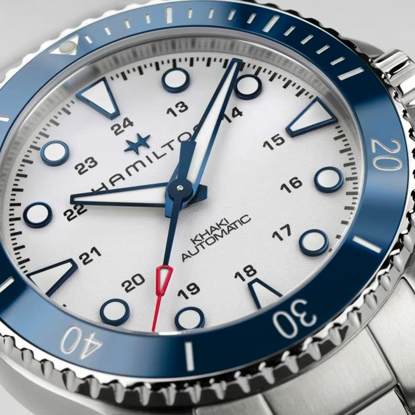 Close-up of a Hamilton Khaki Navy Scuba Auto 43mm watch, capturing the essence of military heritage. It boasts a round, white dial adorned with blue hour markers and numerals for both 12-hour and 24-hour formats, complemented by a blue bezel, silver casing, and a red-tipped second hand.