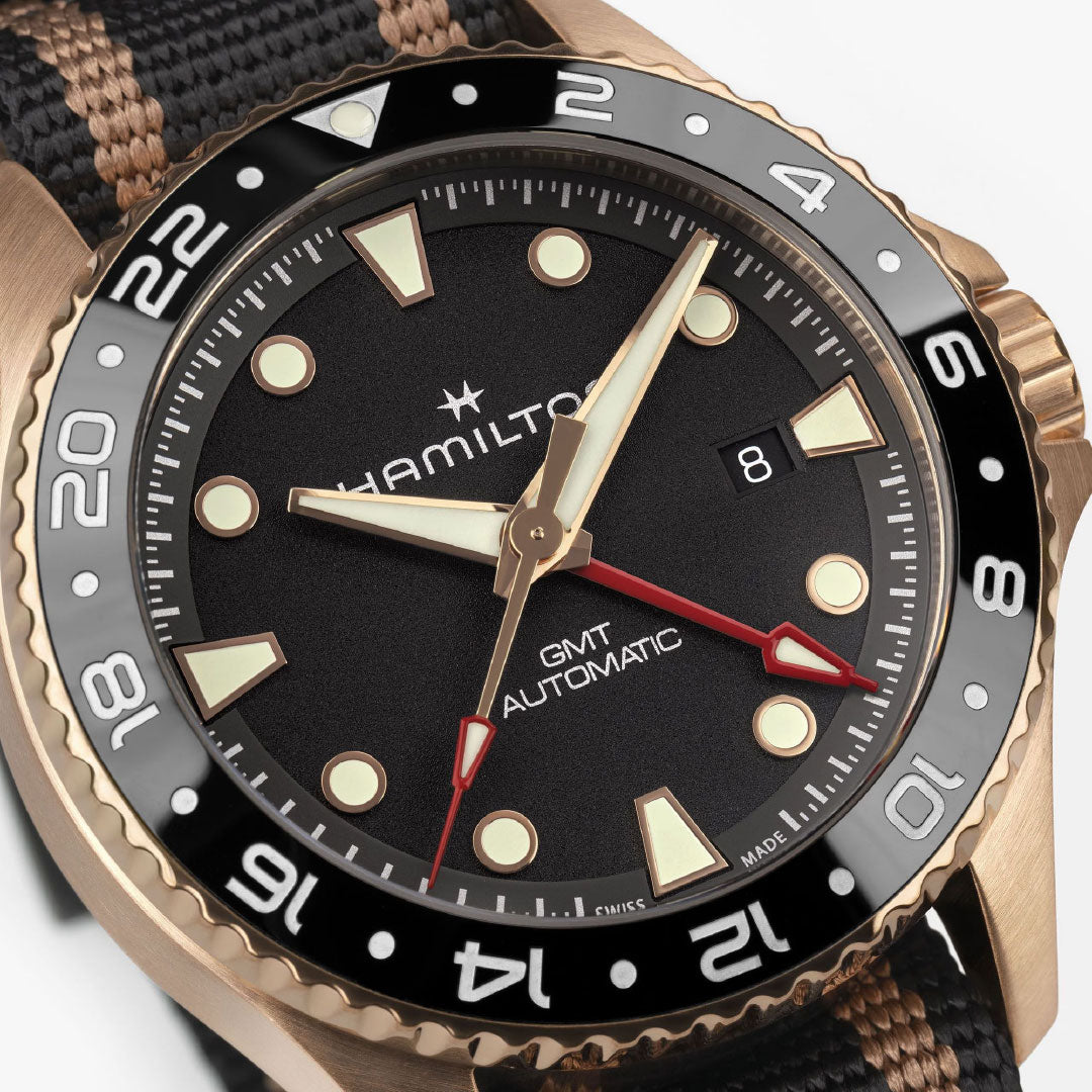 Close-up of the Hamilton Khaki Navy Scuba Automatic GMT 43mm Bronze Watch by Hamilton Watch, featuring a black dial, gold hands and markers, black and bronze bezel, date at 3 o’clock, and a black and tan striped fabric strap.