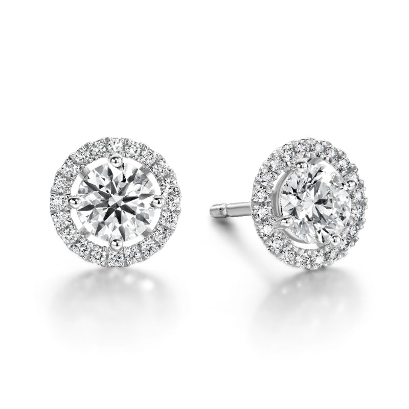 The Hearts On Fire Joy Stud Earrings by Hearts on Fire feature elegant diamond studs, each showcasing a central diamond encircled by a halo of smaller diamonds, all elegantly set in 18 Karat White Gold. These exquisite earrings are displayed side by side against a simple white background.