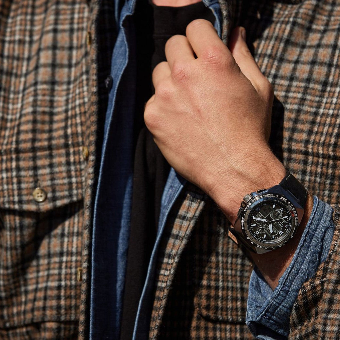A close-up captures an individual in a plaid jacket and denim shirt, adjusting their collar with one hand. On their left wrist is the CITIZEN Promaster Skyhawk A-T Eco-Drive 46mm Watch, its intricate design illuminated as the sun casts shadows to accentuate its details.