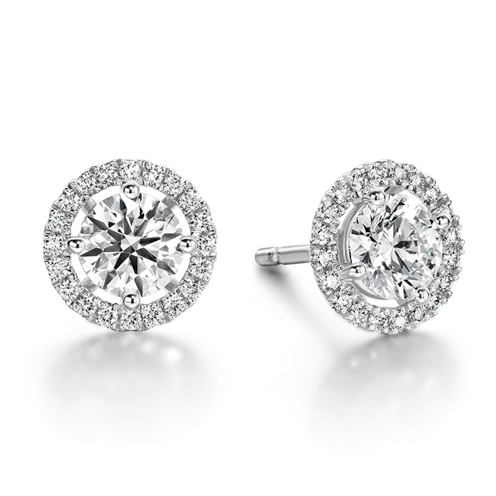 Hearts On Fire Joy 18K White Gold Earrings showcase stunning round diamond studs accented with a halo of smaller diamonds, elegantly set in luxurious white gold and displayed against a pristine white background, crafted with the precision and brilliance synonymous with Hearts on Fire.