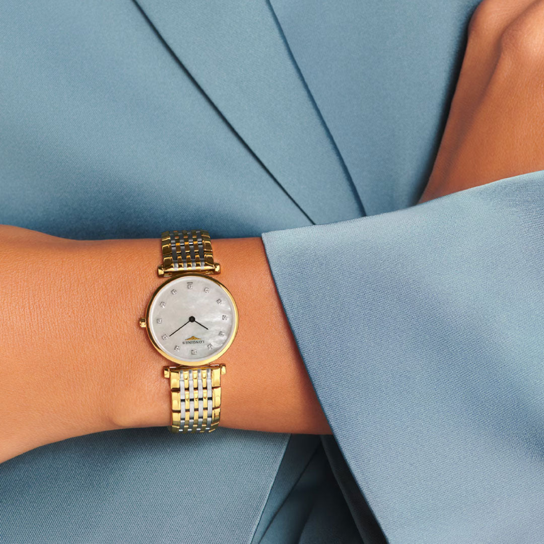A person wearing a Longines La Grande Classique Quartz 24mm gold and silver stainless steel watch with a white round face, diamond hour markers, scratch-resistant features, and dressed in a light blue long-sleeve garment.