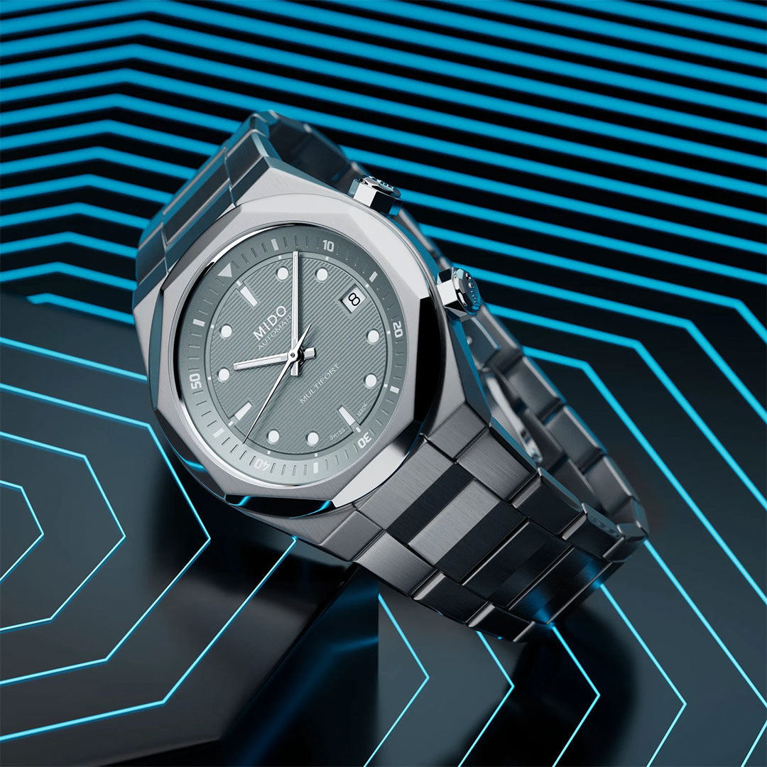 The MIDO Multifort 8 Two Crowns Automatic 40mm watch is shown at an angle on a metallic strap, featuring a geometric-patterned dial and set against a blue and black neon hexagonal background.