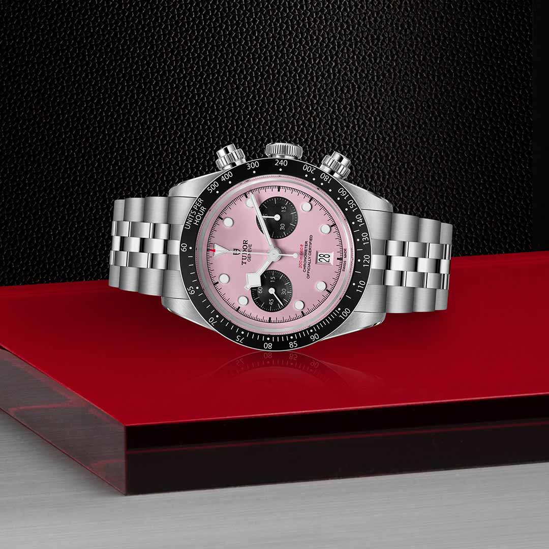 The TUDOR Black Bay Chrono by TUDOR features a steel case, silver bracelet, black bezel, pink dial, and two black subdials, displayed on a reflective red and silver surface against a textured black background.