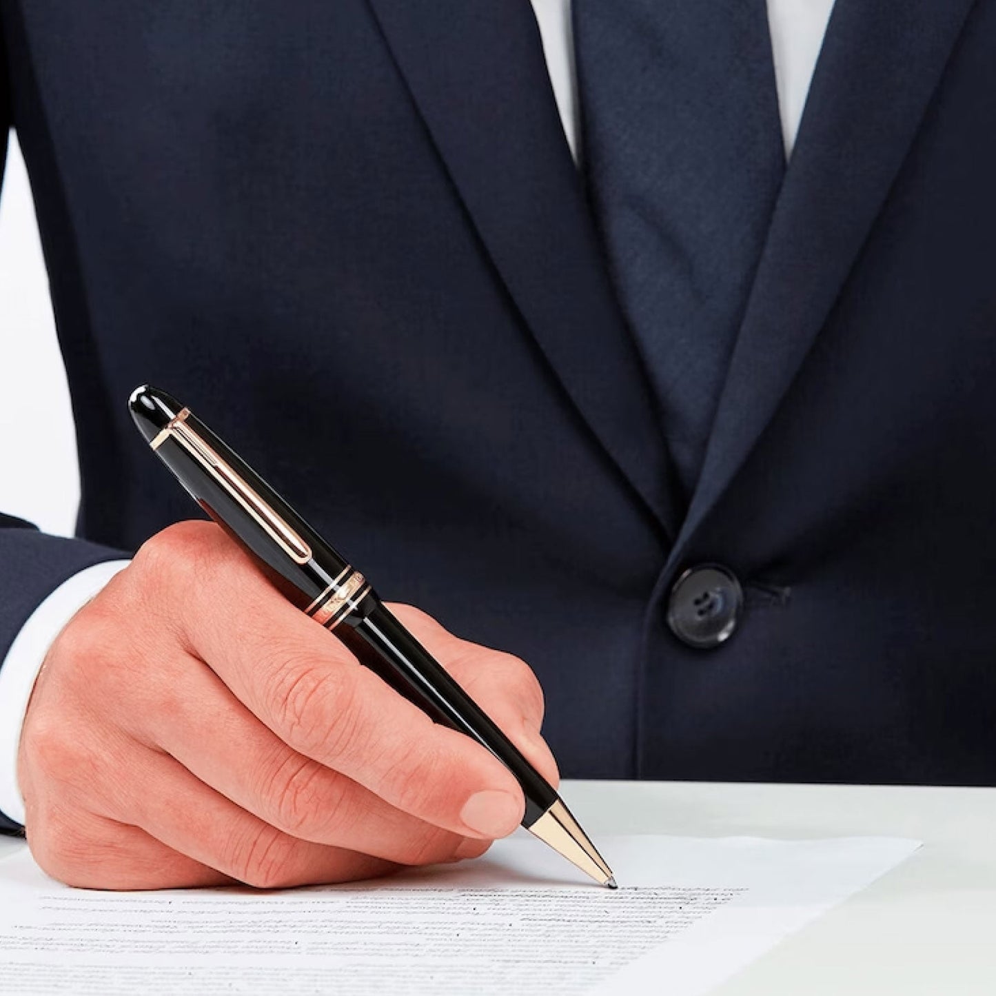 A person in a navy suit holds a Montblanc Meisterstück Black Resin Rose Plated Ballpoint Pen, poised above a document on a white table. The focus is on the luxury writing instrument and hand, suggesting the action of signing or writing.