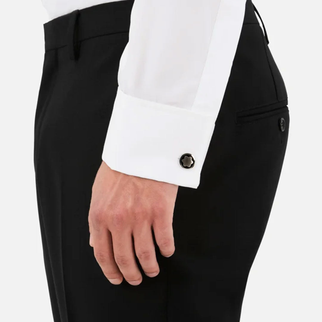 A close-up of a person wearing a white dress shirt with black pants features the Montblanc Star Black Onyx and Stainless Steel Cufflinks adorning the shirt cuff, with the hand relaxed at the side. The background is plain and light-colored.