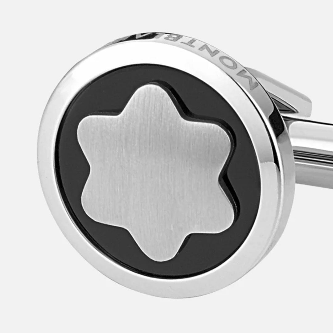 A close-up of a Montblanc Star Black Onyx and Stainless Steel Cufflink showcases its stainless steel finish with the iconic six-pointed snowcap emblem set against a black circular background. The metal border, engraved with the Montblanc signature, beautifully frames this exquisite design.