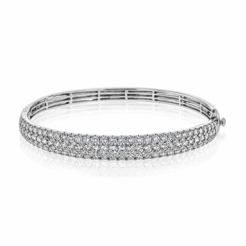 The Simon G. 18K White Gold Three Row Diamond Bangle, elegantly designed by Simon G, features a graceful circular shape with multiple rows of dazzling white diamonds set in 18k white gold. Its reflective finish adds to its luxurious appeal as it rests against a plain white background.