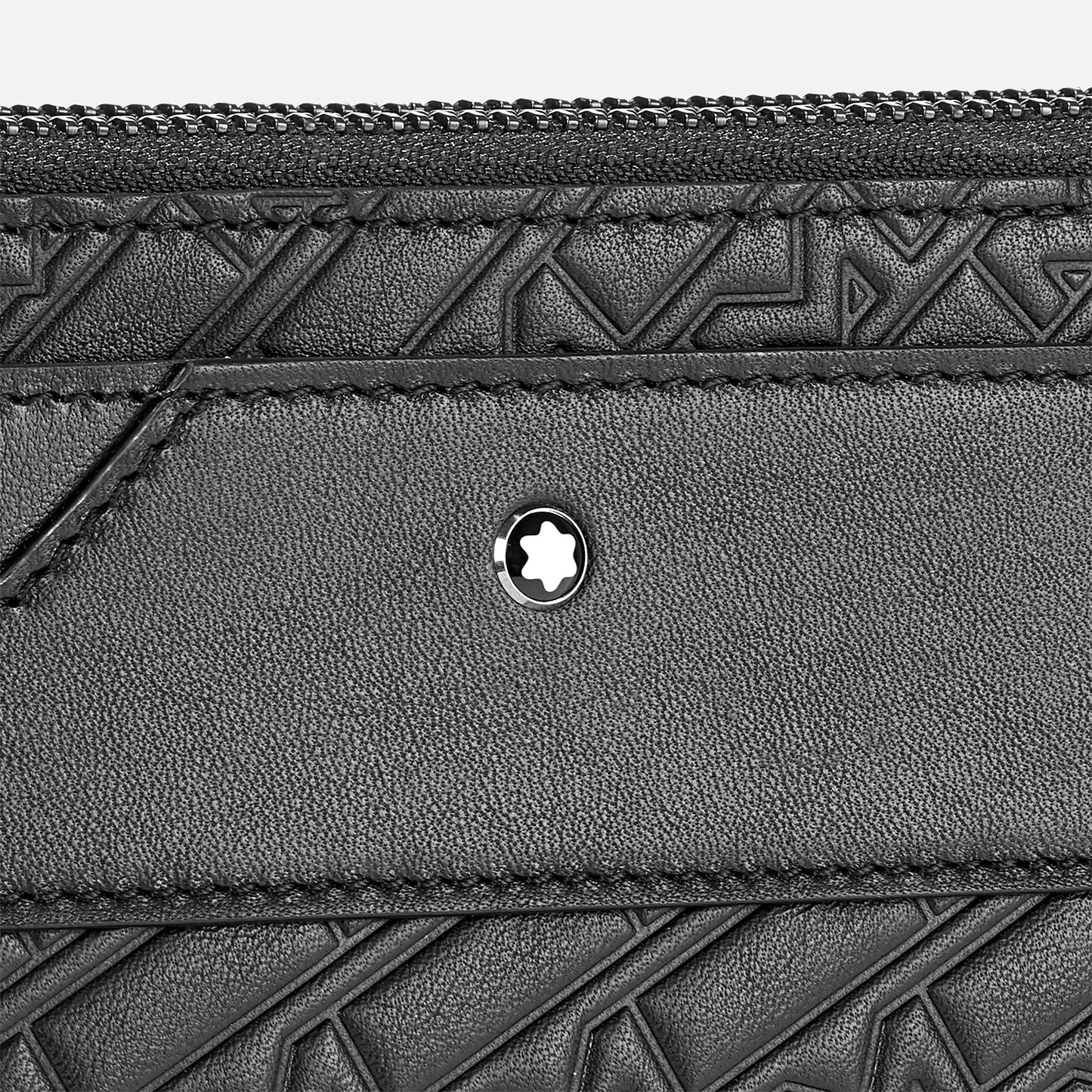 A close-up of the Montblanc M-Gram Leather Clutch Bag showcases its black textured leather with a geometric embossed pattern. Centered on the leather is a circular emblem featuring a star-like design. At the top, the stylish Montblanc M-Gram 4810 zipper is prominently visible.
