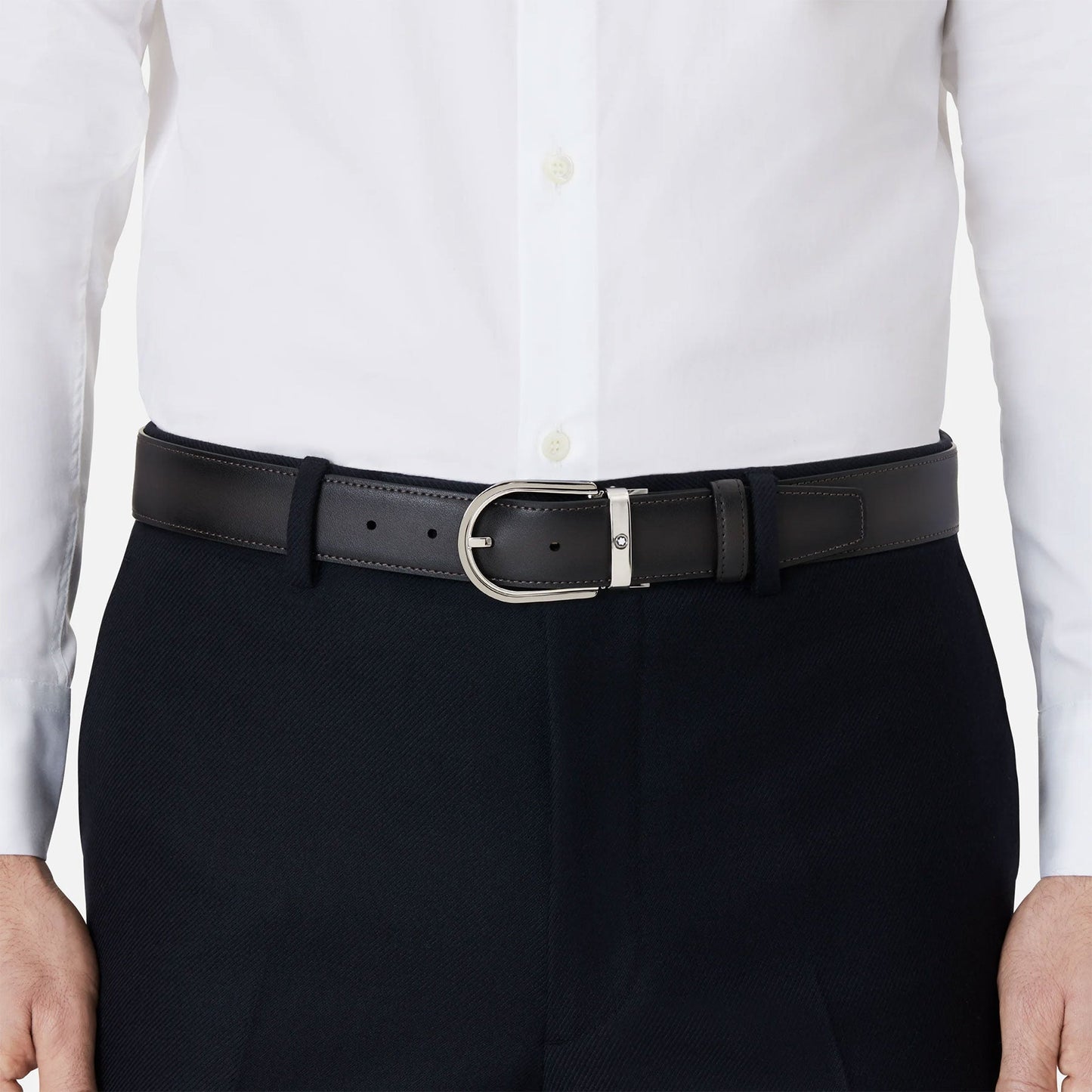 A person wearing a white dress shirt and black formal pants, enhanced by the Montblanc Horseshoe Leather Belt, with its unique horseshoe buckle. The shirt is neatly tucked into the pants.