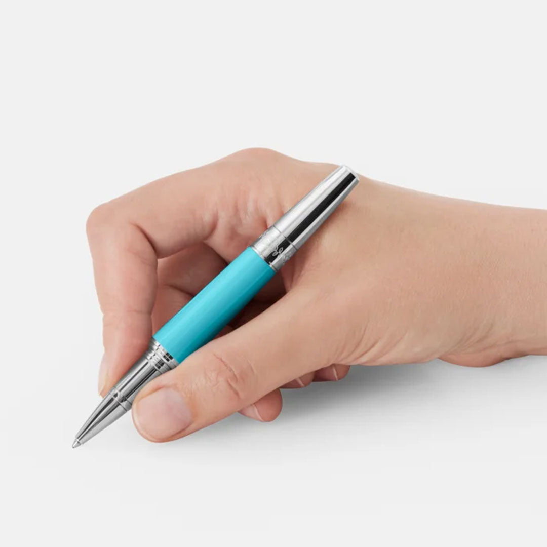A hand delicately grasps a Montblanc Muses Maria Callas Special Edition Rollerball Pen, showcasing its elegant silver and turquoise design against a simple white backdrop. The pen is held in position, appearing ready for use.