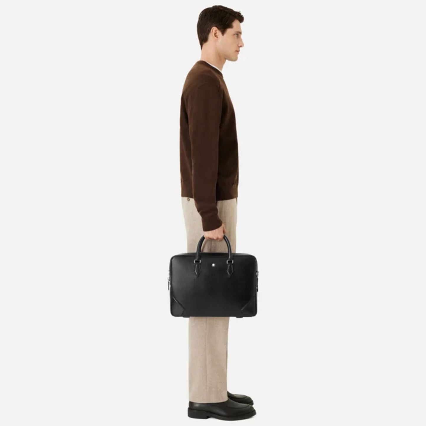 A person in a brown sweater and beige pants stands in profile against a plain background, holding a Montblanc Meisterstück Black Leather Document Case by their side.