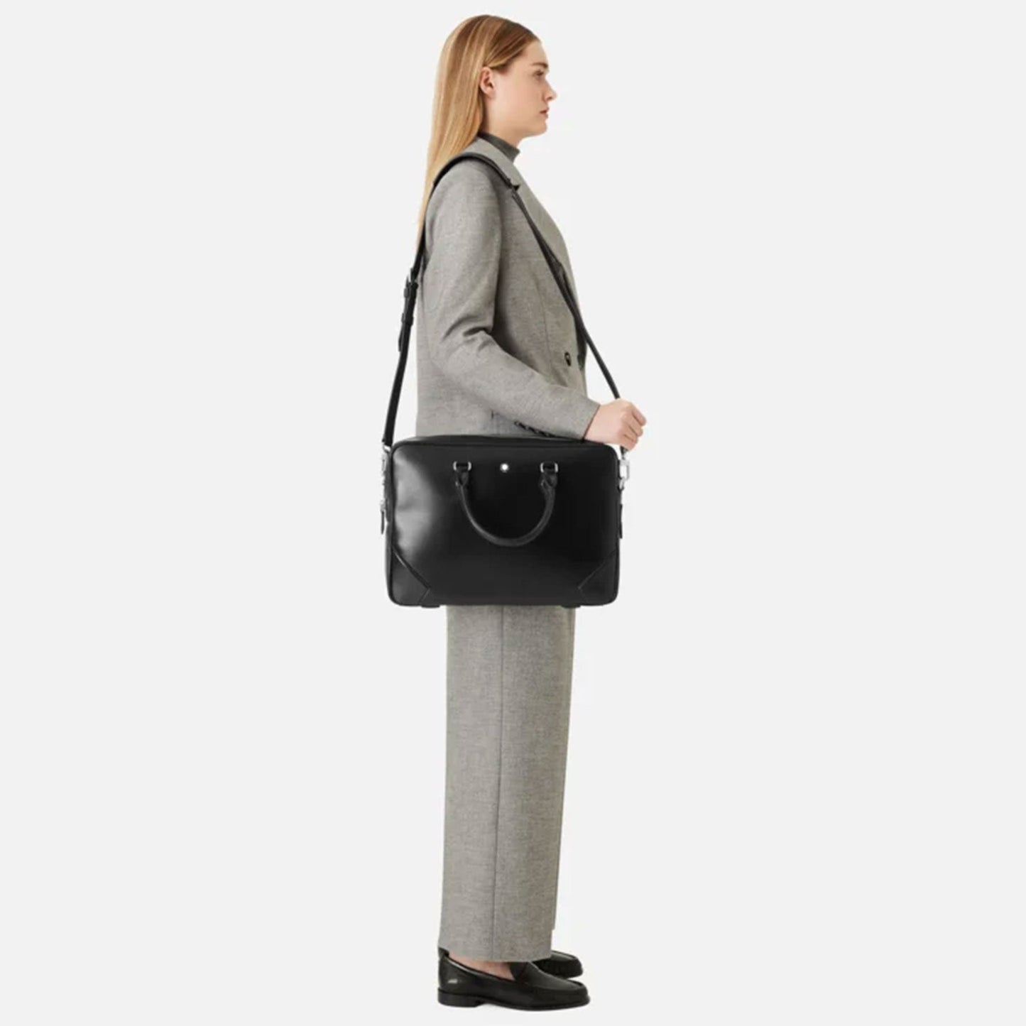 A person with long hair, dressed in a gray business suit and black loafers, stands sideways holding a Montblanc Meisterstück Black Leather Document Case over their shoulder. The background is plain white, perfectly accentuating this timeless style from Montblanc.