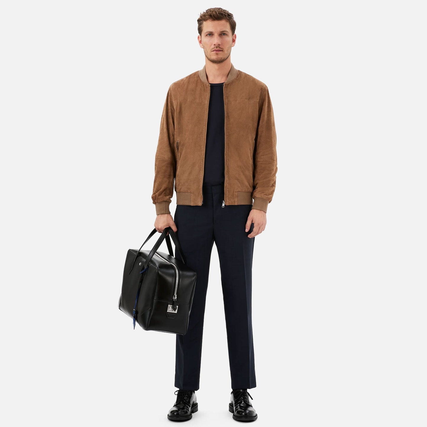 A man stands against a plain background, wearing a brown suede jacket, black shirt, and dark trousers. He holds a Montblanc Meisterstuck Leather Duffle Bag in his left hand and wears black shoes.