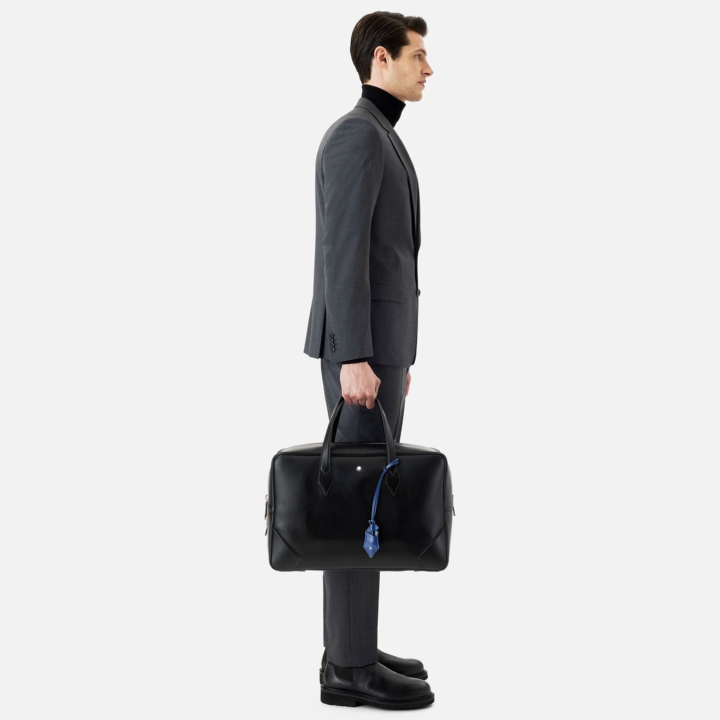 A man in a gray suit and black turtleneck stands in profile, holding a Montblanc Meisterstuck Leather Duffle Bag distinguished by its striking blue tassel. He completes the ensemble with sleek black shoes against a plain white background.