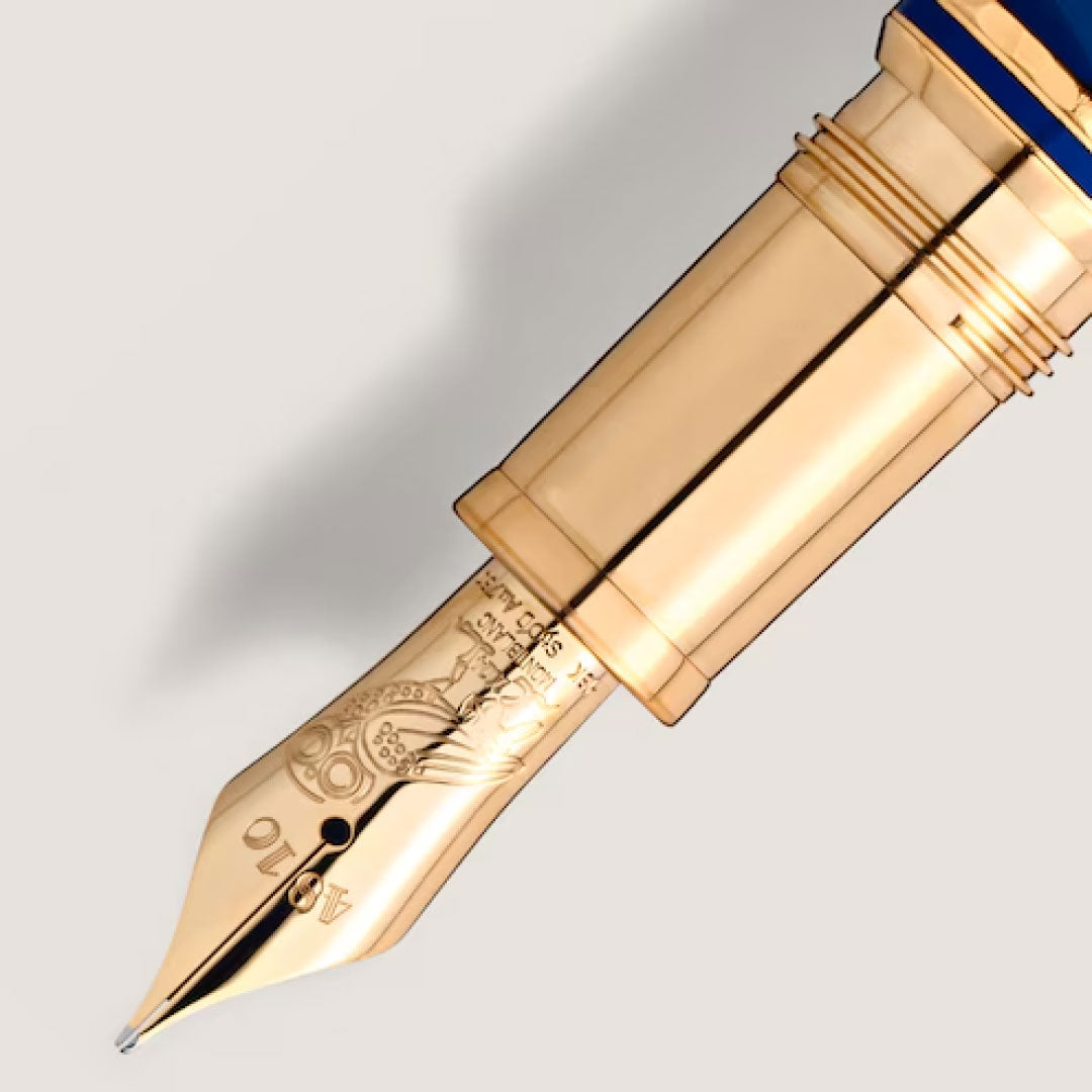 A close-up shot features the gold nib of the Montblanc Masters Of Art Homage To Gustav Klimt Limited Edition Fountain Pen, adorned with intricate engravings that echo Gustav Klimt's artistic detailing. The partially visible barrel exhibits a glossy blue finish, exemplifying Montblanc's fine craftsmanship against a soft, off-white background.