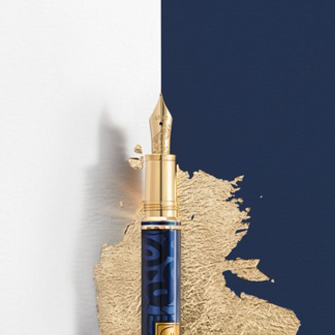 The Montblanc Masters Of Art Homage To Gustav Klimt Limited Edition Fountain Pen, featuring a gold nib and blue barrel, is placed upright against a backdrop of white and navy blue. Gold paint splashes around it in an artistic manner that captures the essence of the Vienna Secession, reminiscent of Klimt's unique style.