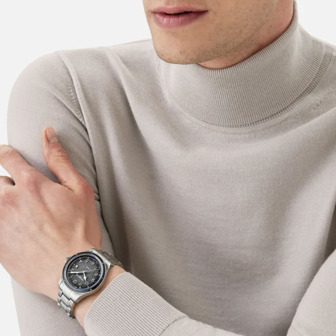 A person wearing a light gray turtleneck sweater and a distinguished Montblanc 1858 Date 0 Oxygen The 8000 Automatic 41mm Watch with a dark face crosses their arms, revealing only the lower half of the face and upper torso. The focus is on the watch, a symbol of precision that embodies the spirit of exploration.