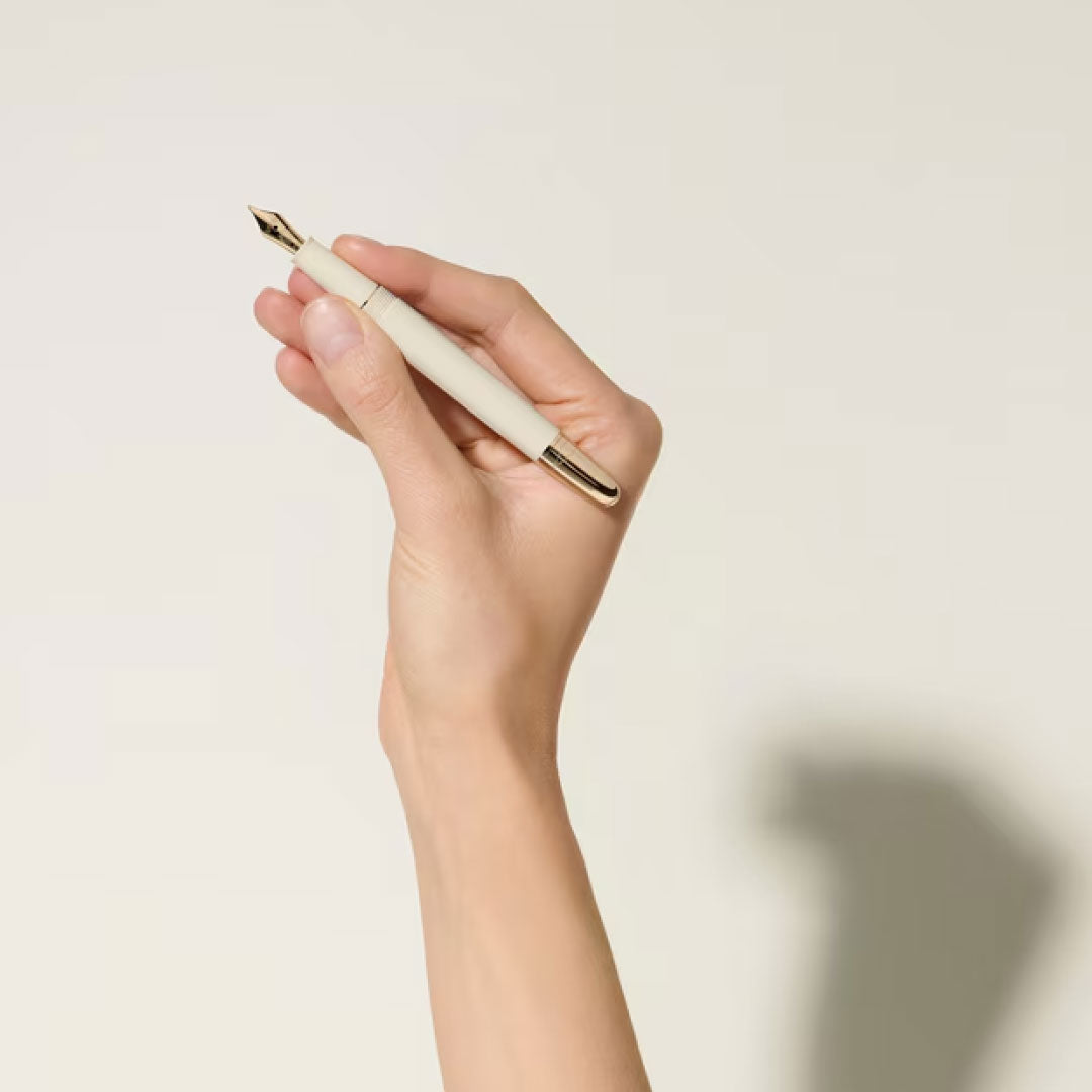 A hand holds a Montblanc Meisterstuck Romeo & Juliet Doue Classique Ballpoint Pen upright against an off-white background, its tip pointing upward and casting a gentle shadow on the wall.