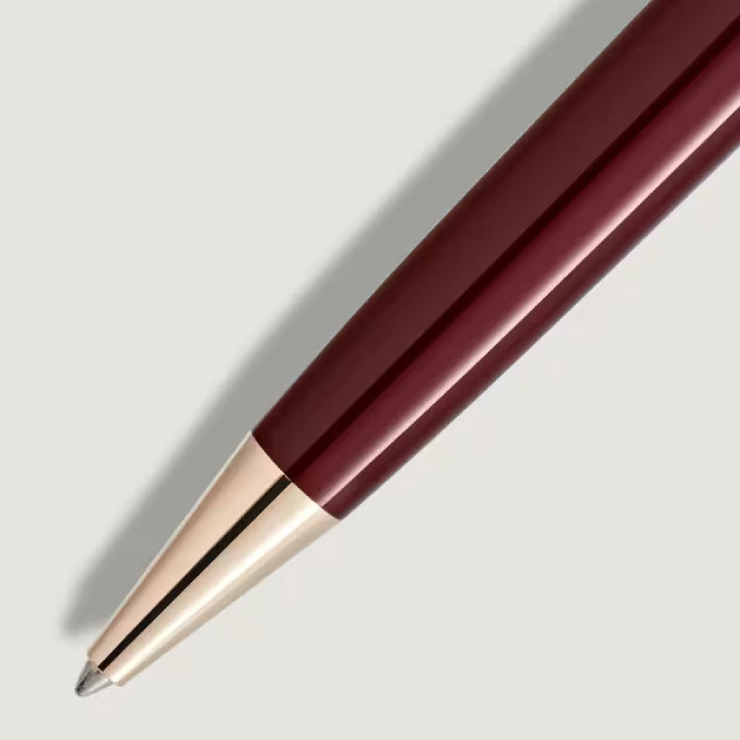 A close-up of the Montblanc Meisterstuck Burgundy Midsize Ballpoint Pen, accented with gold fittings, shows its burgundy and gold tip resting on a light gray surface and casting a soft shadow to the left.