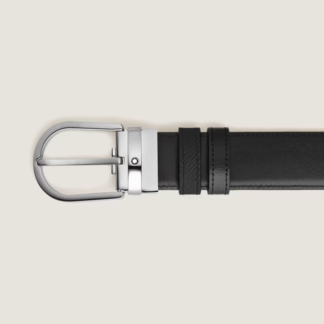 A close-up of the Montblanc Horseshoe Buckle 35mm Reversible Leather Belt in black saffiano leather with a polished silver buckle featuring a circular emblem, displayed horizontally on a light beige background.