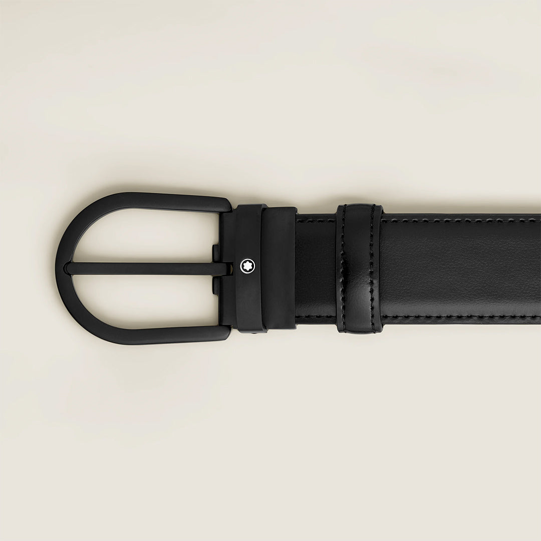 The Montblanc Horseshoe Buckle 35mm Black Leather Belt features a matte black rounded buckle with a subtle circular logo by the hole, set against a light beige background.