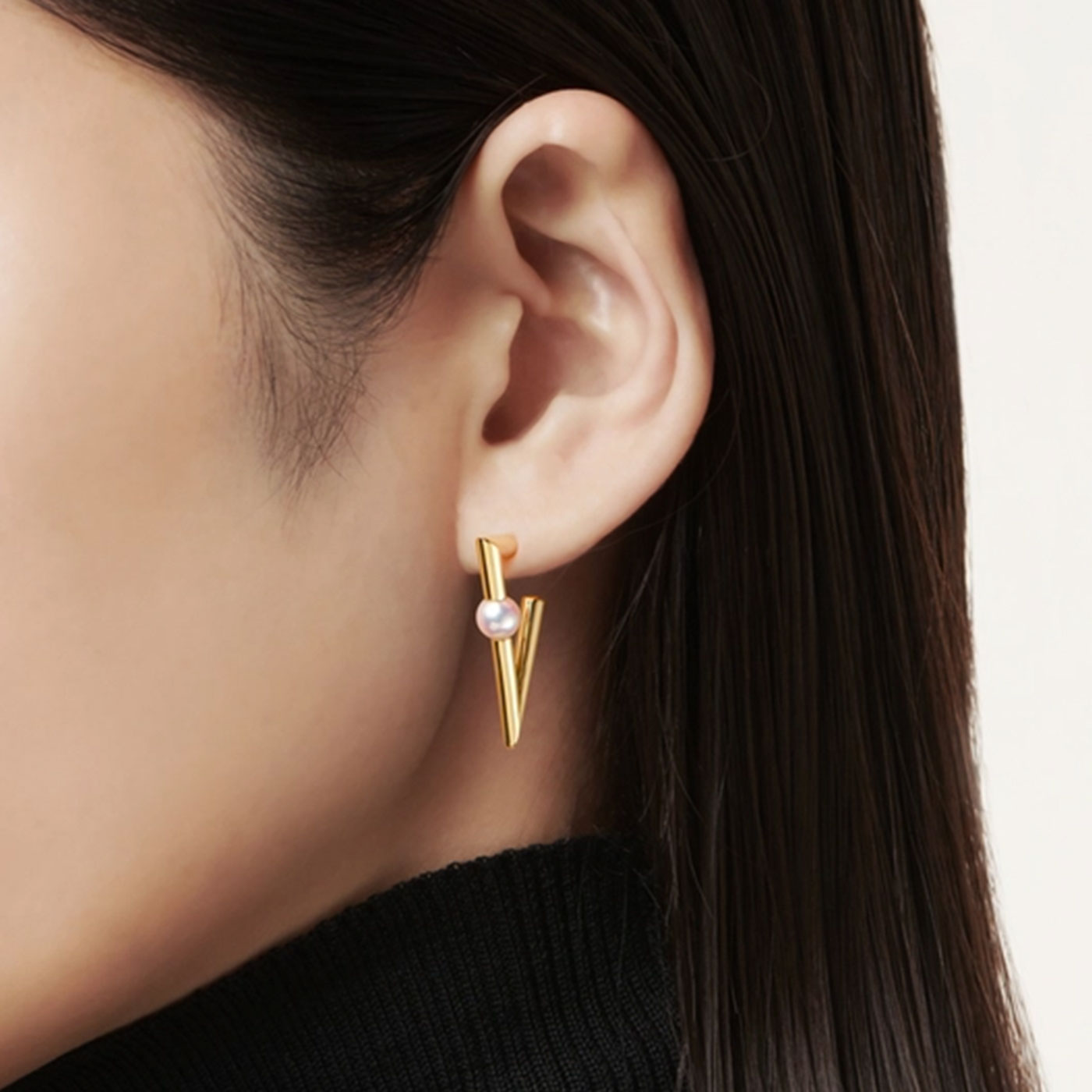 The photograph captures a person with straight, dark brown hair wearing a black turtleneck sweater. Their ear is adorned with the Mikimoto V Code 18K Yellow Gold Pearl Hoop Earring, showcasing its triangular design and Akoya cultured pearl accent.