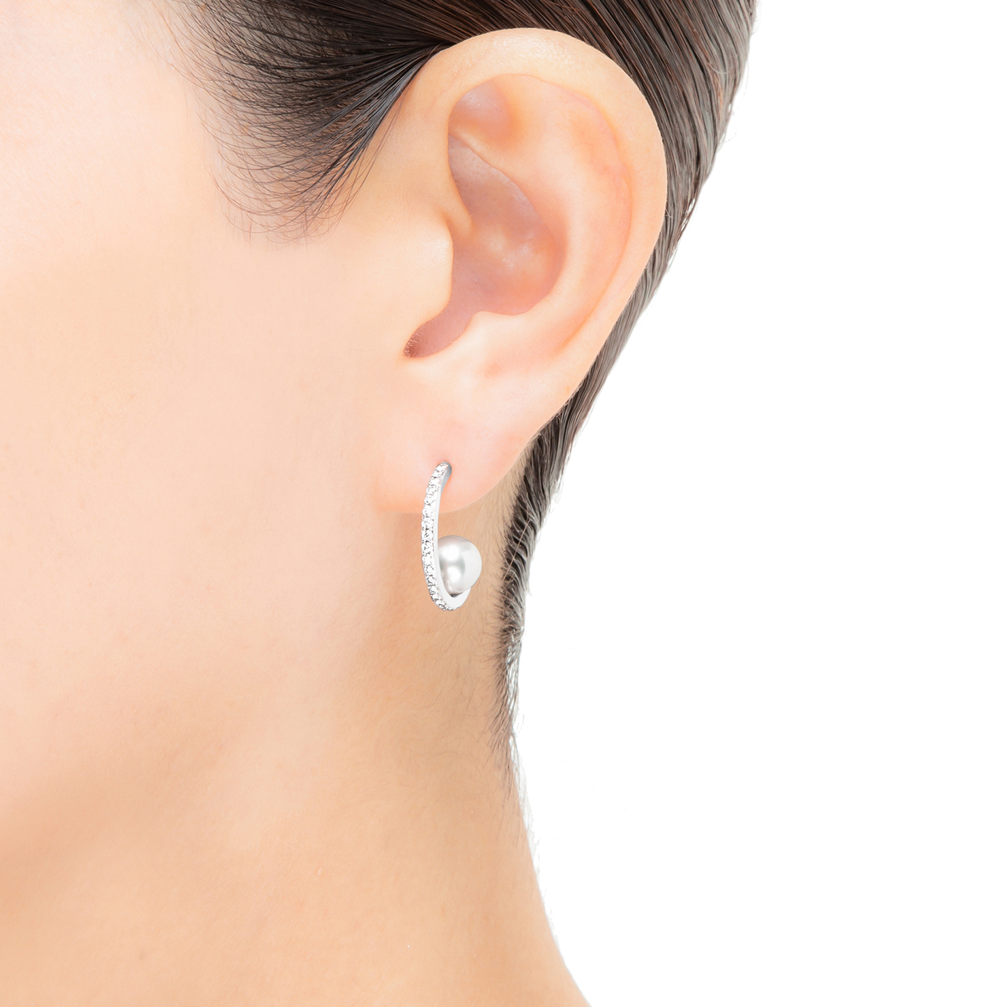 A close-up of a person wearing Mikimoto 18k White Gold Akoya Cultured Pearl and Diamond Hoop Earrings showcases their elegant design. The silver-tone earring, featuring a curved, sparkling appearance adorned with cultured pearls, rests gracefully on the earlobe against a white background. With the hair pulled back, only the ear and part of the neck are visible.