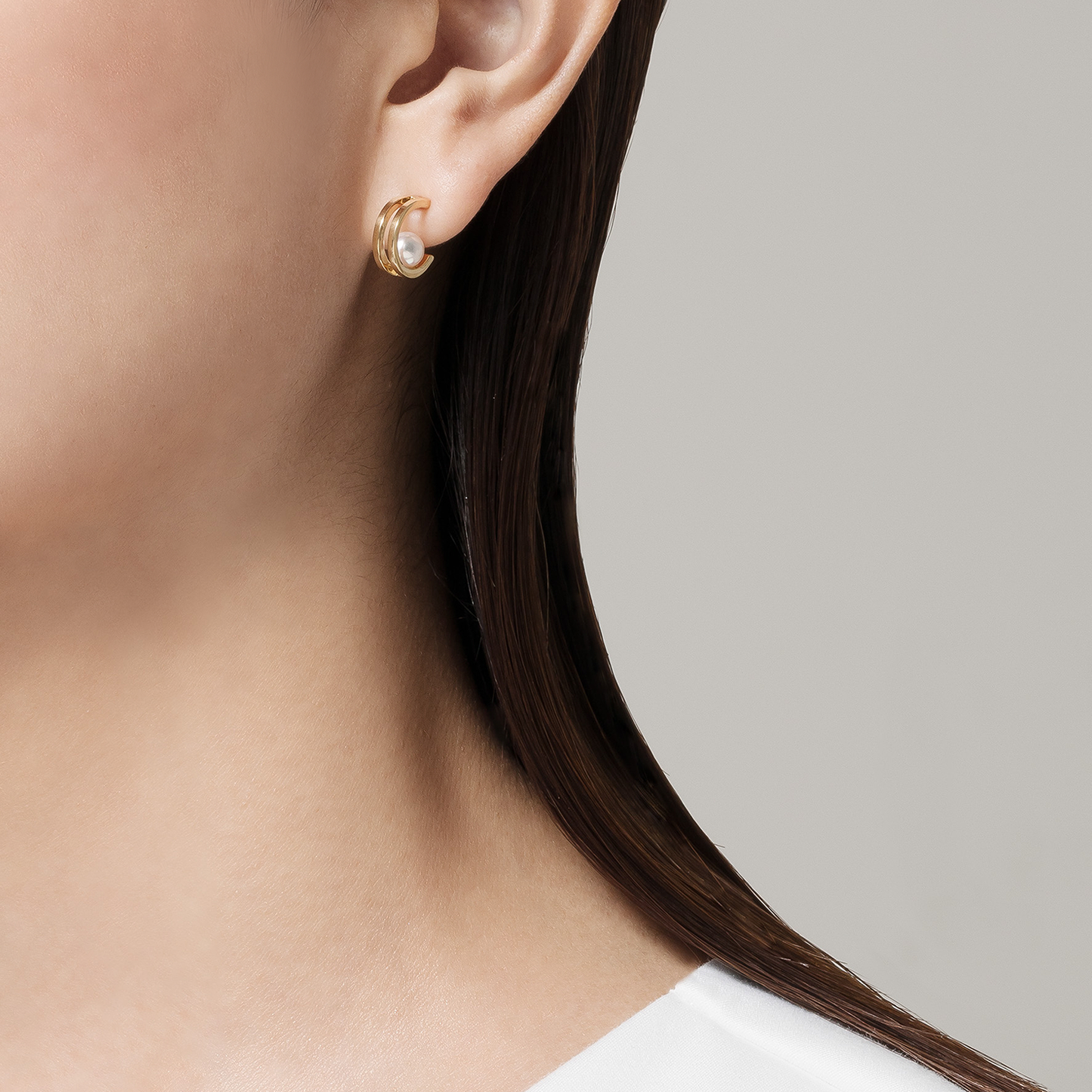 A close-up of a woman's ear showcases a Mikimoto 18K Yellow Gold Akoya Cultured Pearl Hoop Earring. The background is plain light gray, and she is dressed in a white shirt.