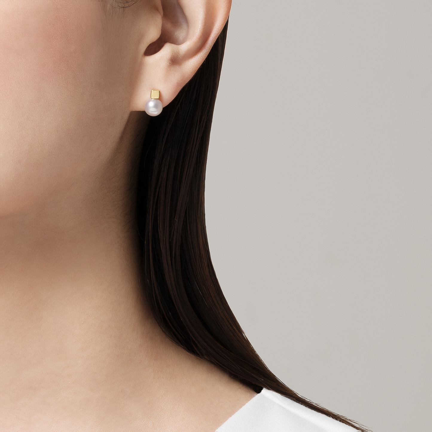 In the image, a close-up reveals a person's ear graced by Mikimoto's Akoya Cultured Pearl Stud Earring, enhanced with an 18K yellow gold accent. The individual has straight, dark hair and is dressed in a white top, set against a neutral gray background.