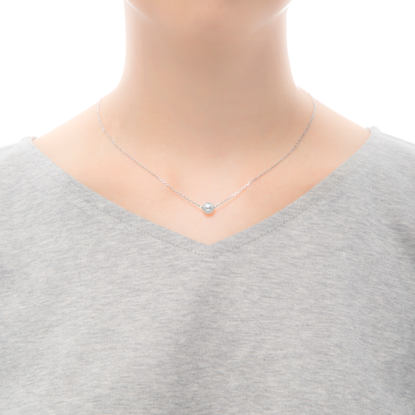 A person wearing a plain gray V-neck shirt is shown. They sport the Mikimoto 18k White Gold Akoya Cultured Pearl Pendant, which features a delicate silver necklace adorned with an Akoya cultured pearl at its center. The background is white.