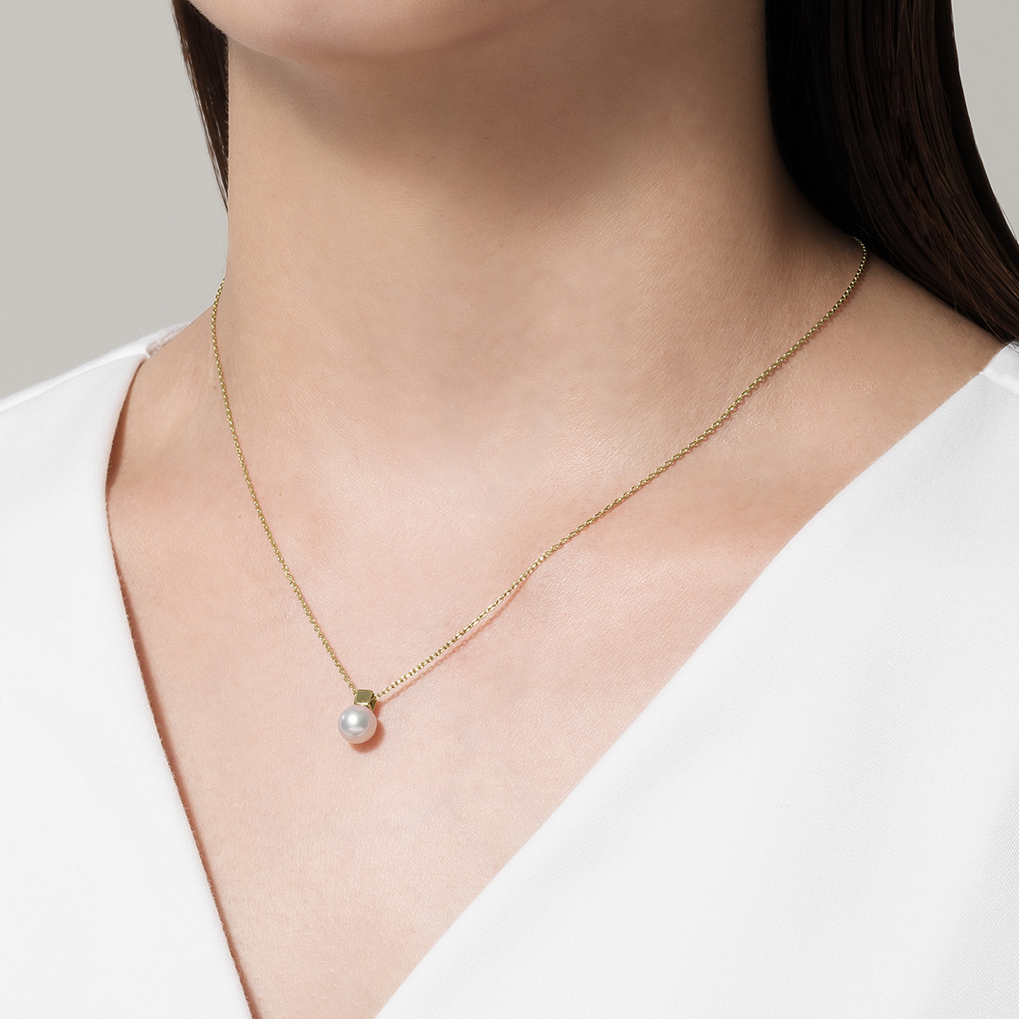 A person is wearing a Mikimoto 18K Yellow Gold Akoya Cultured Pearl Necklace Pendant. They have straight, brown hair and are dressed in a white V-neck top, with the yellow gold chain adding an elegant touch against the neutral background.