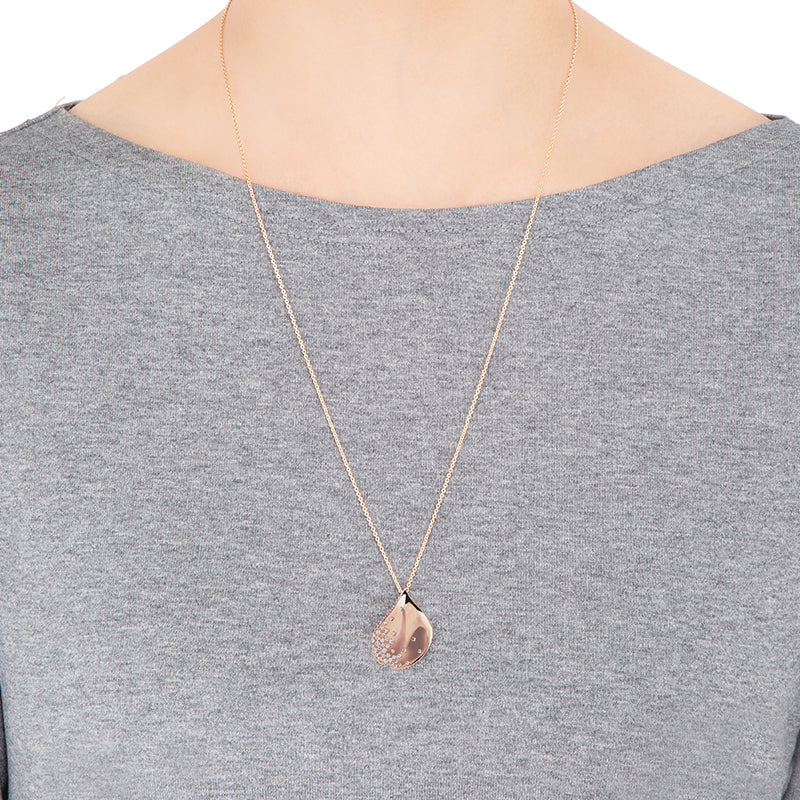 A person is wearing a gray top and a long necklace featuring the Mikimoto Les Pétales de Ginza 18K Rose Gold Diamond Necklace Pendant. The shiny pendant, in the form of a teardrop, hangs at chest level.
