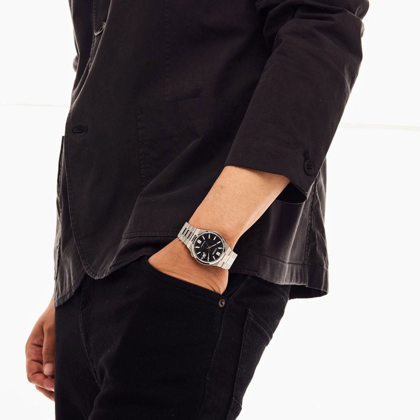 A person dressed in a black jacket and pants stands casually with their left hand in their pocket. On their wrist is a CITIZEN TSUYOSA Collection Automatic Mechanical 40mm Watch, featuring sports styling with a silver finish and a black face, offering water resistance. The background is white and minimal.