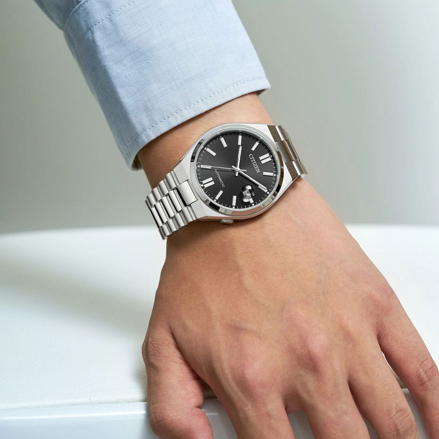 An individual is wearing a CITIZEN TSUYOSA Collection Automatic Mechanical 40mm Watch, featuring a silver wristband and black dial with Roman numerals and a date window. This automatic watch by CITIZEN rests on a light-colored surface, complemented by a light blue long-sleeve shirt.