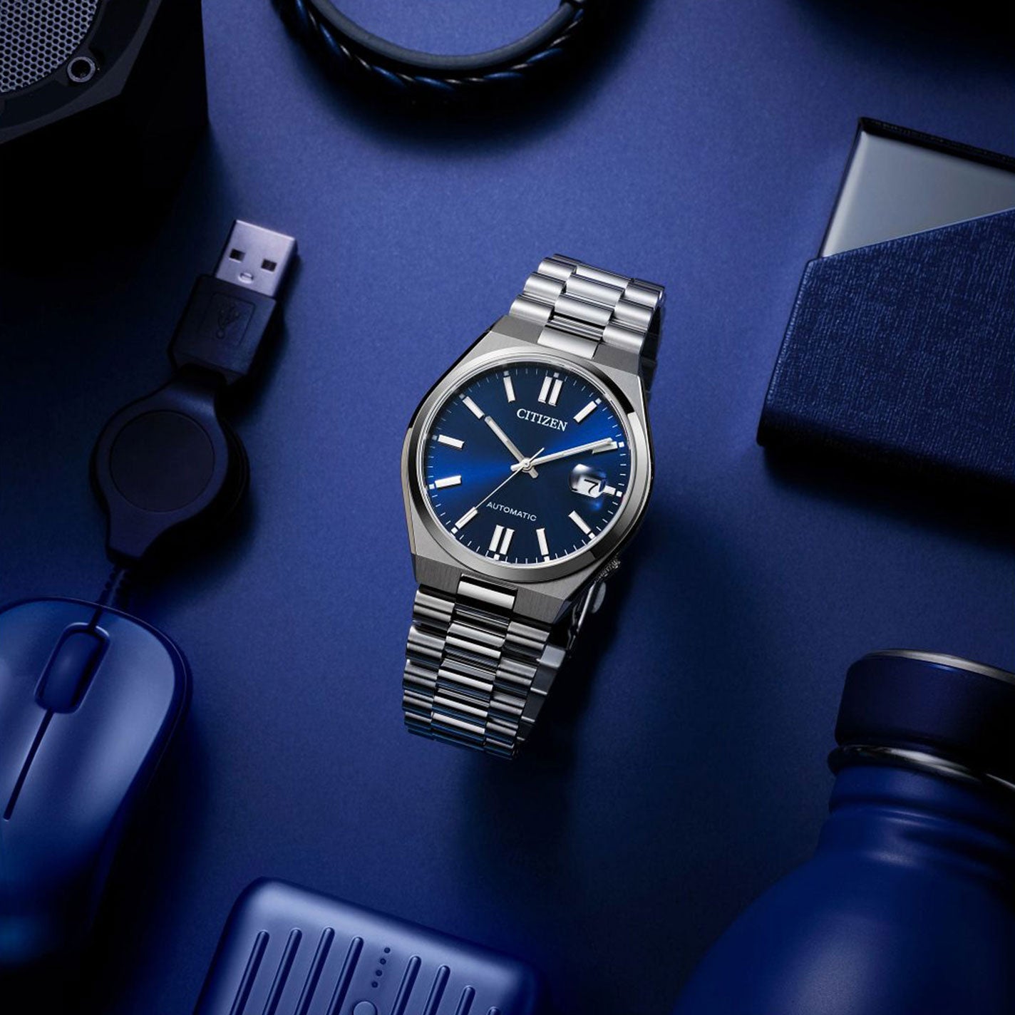 A CITIZEN TSUYOSA Collection Automatic Mechanical 40mm watch, characterized by its silver design with a blue face and silver numerals, rests on a blue surface. The sleek sports styling harmonizes with nearby items such as a water bottle, USB cable, computer mouse, and small case—all in shades of blue.