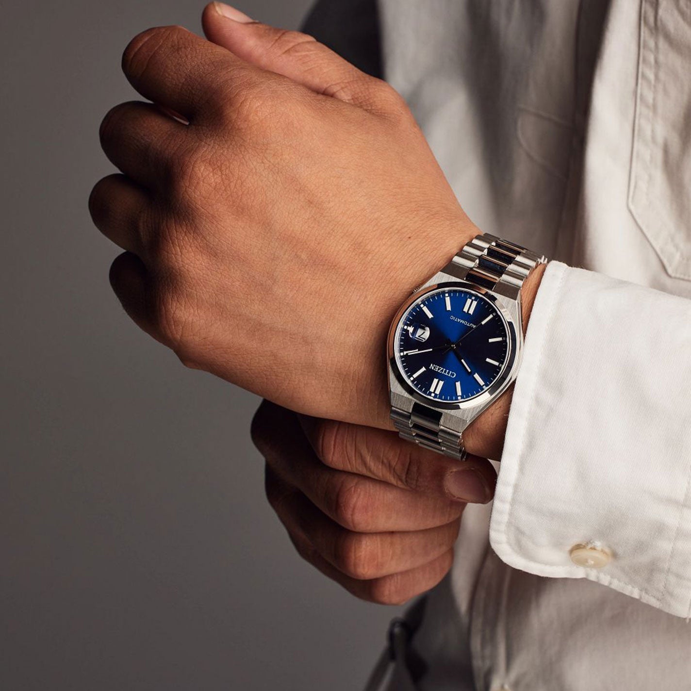 The image zooms in on a person wearing a CITIZEN TSUYOSA Collection Automatic Mechanical 40mm Watch, showcasing its ultramodern sports styling with a silver finish and blue dial, complete with a date display. The watch adorns their left wrist as they adjust it with their right hand while dressed in a white shirt against a neutral background.