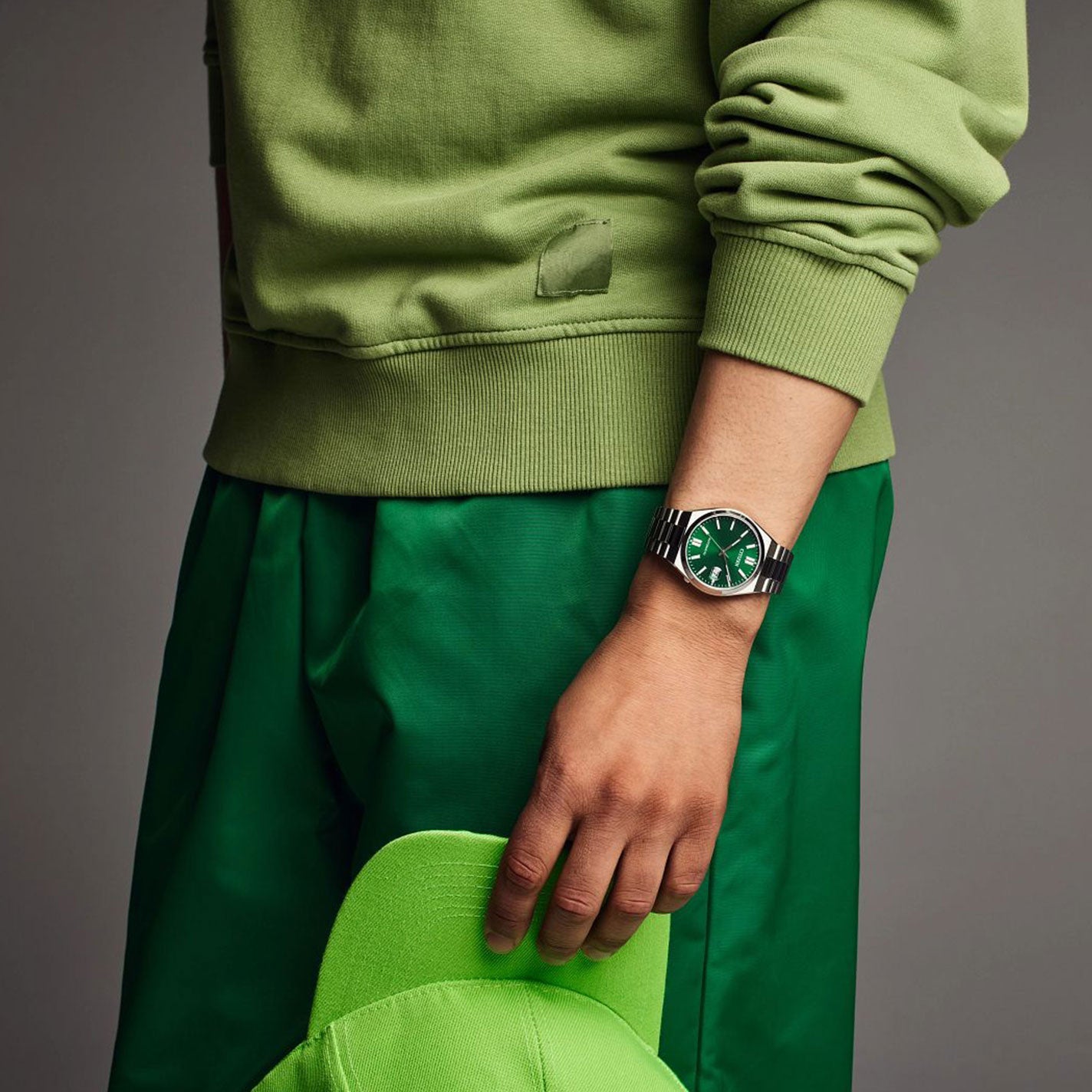 A person in a green hoodie and matching pants holds a cap, their wrist adorned with the CITIZEN TSUYOSA Collection Automatic Mechanical 40mm Watch, which boasts an ultramodern sports styling and sunray green dial. The neutral gray background adds contrast to their vibrant ensemble.
