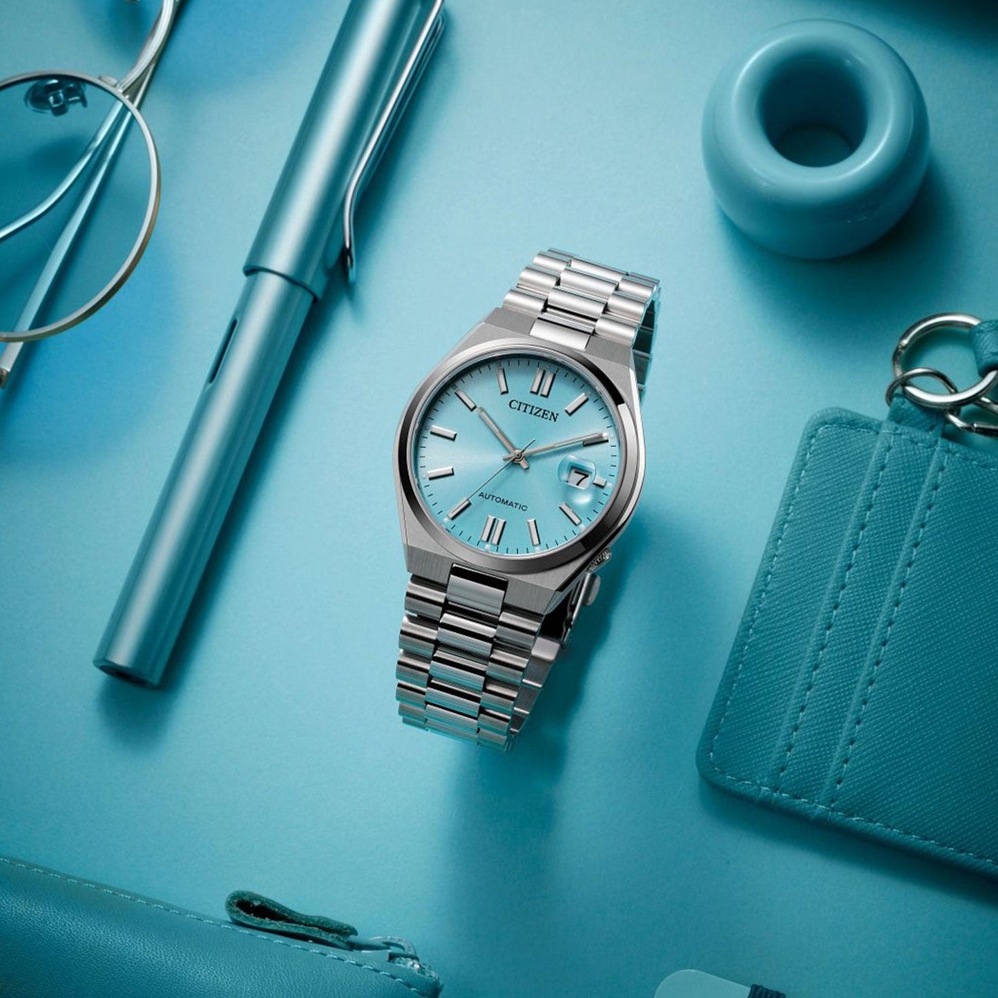 A CITIZEN TSUYOSA Collection Automatic Mechanical 40mm Watch, featuring a teal dial and stainless steel case, is placed on a blue background. Surrounding items include a pen, ring, glasses, card holder, and small pouch, all in shades of blue.