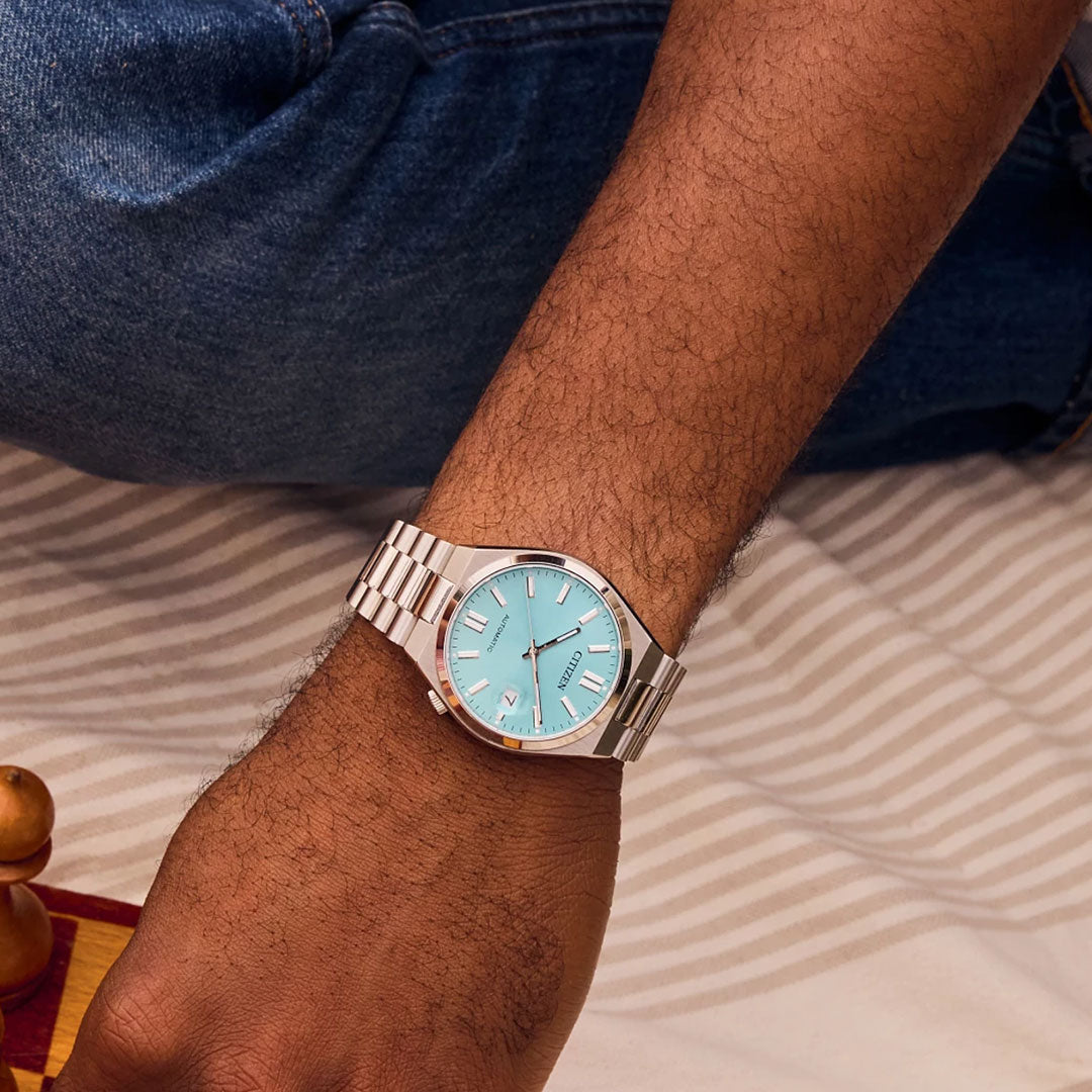 A person wearing the CITIZEN TSUYOSA Collection Automatic 37mm Watch with a sunray-textured light blue dial and silver markers rests their arm on a striped surface, the watch face contrasting stylishly with blue jeans.