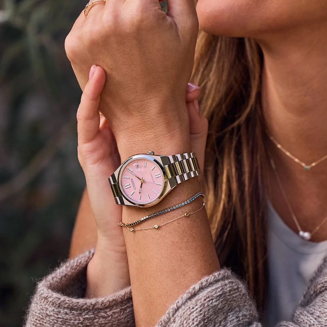 A woman wears a CITIZEN TSUYOSA Collection Automatic 37mm Watch by CITIZEN, featuring a pastel pink dial and stainless steel case, styled with gold bracelets and a necklace. Her hands rest on her sweater, her face partially visible.