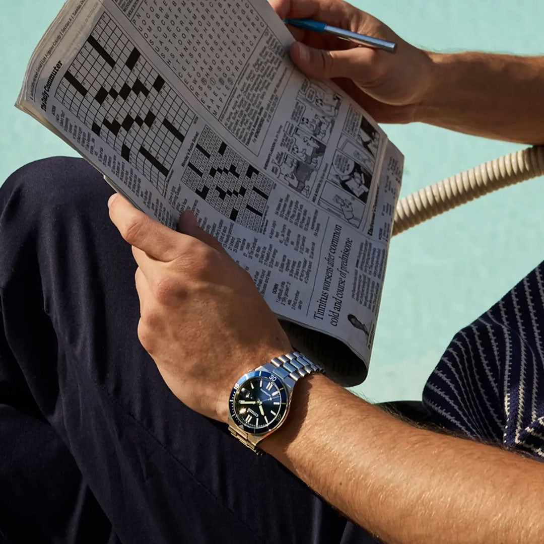 A person by a pool solves a newspaper crossword while wearing the CITIZEN Tsuyosa Shore Automatic 40mm Watch.