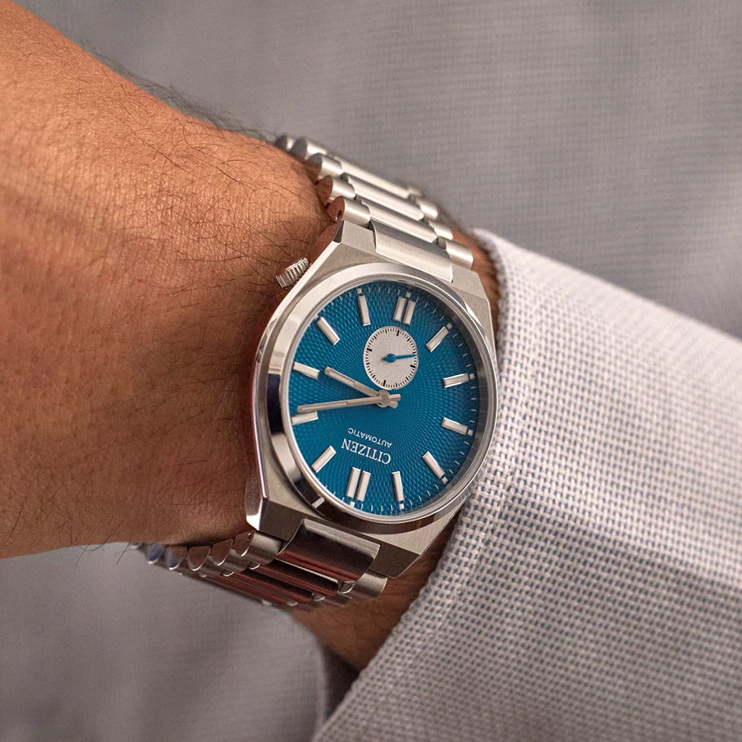 A person wearing a CITIZEN TSUYOSA Small Second Automatic 40mm watch with a silver metal bracelet, blue dial, sapphire crystal, silver hour markers, and subdial at 12 o’clock, paired with a light checkered shirt.
