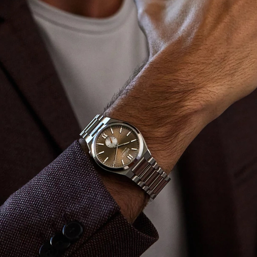 A person in a brown suit jacket and white shirt displays the CITIZEN TSUYOSA Small Second Automatic 40mm Watch by CITIZEN, featuring a green dial and stainless steel case, on their wrist.