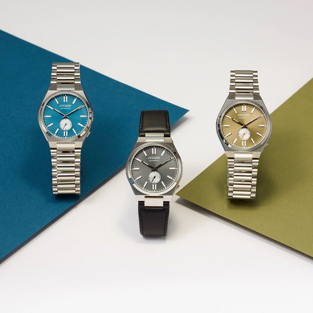 Three CITIZEN TSUYOSA Small Second Automatic 40mm Watches are shown: one with a blue face and metal band, one with a gray face and black leather band, and one with a gold face and metal band, all arranged on colorful paper backgrounds.