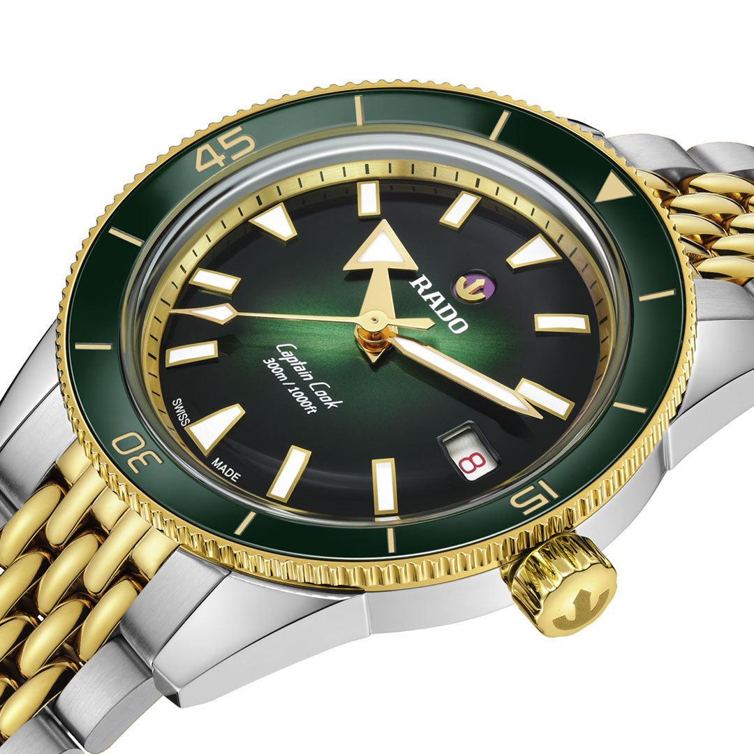 A close-up of the RADO Captain Cook Automatic 39mm Watch featuring a green bezel, gold accents, black gradient dial, two-tone gold and silver bracelet, automatic movement, date at 3 o'clock, and luminous hands and markers.