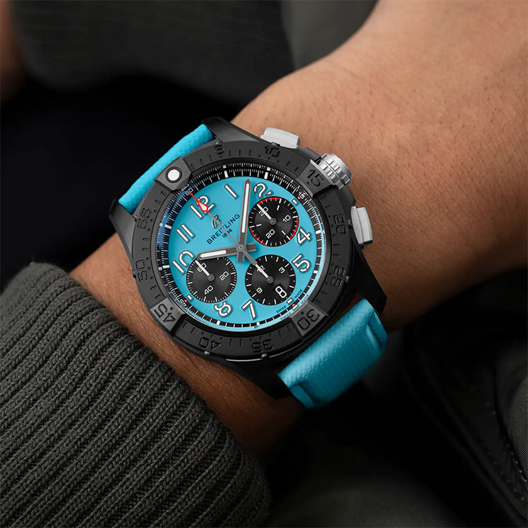 A person wears a Breitling Avenger B01 Chronograph 42 Night Mission with a bright turquoise dial and matching leather strap, paired with a dark, textured sleeve.